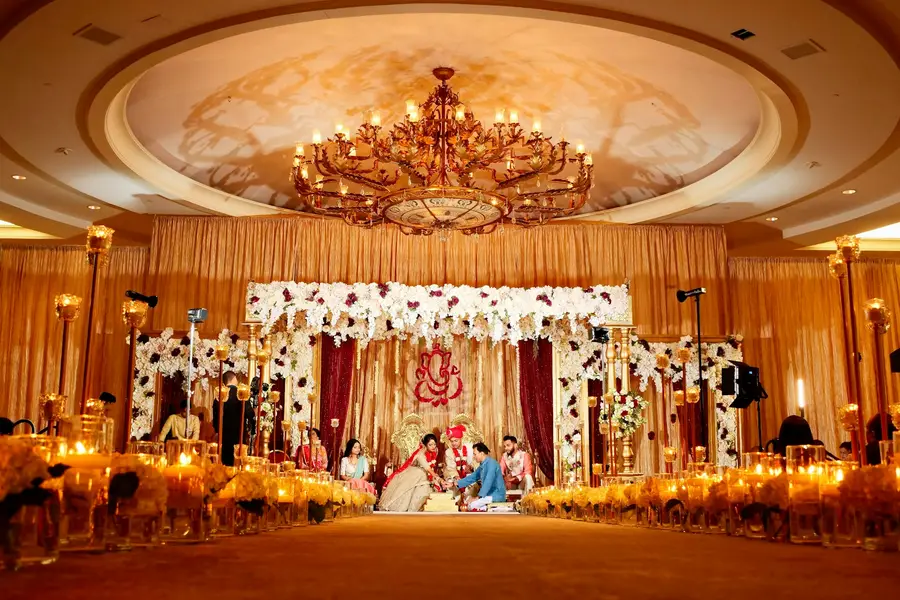 A couple perform a wedding ritual beneath a chandelier in a candlelit ballroom